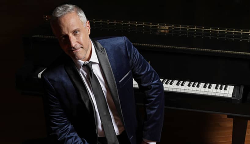 A photo of pianist Jim Brickman sitting in front of a piano wearing a navy suit. 