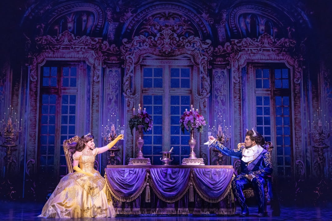 Kyra Belle Johnson & Fergie L. Philippe in beautiful costumes toast each other at a dinner table in Disney's Beauty And The Beast. Photo By Matthew Murphy. (C) Disney